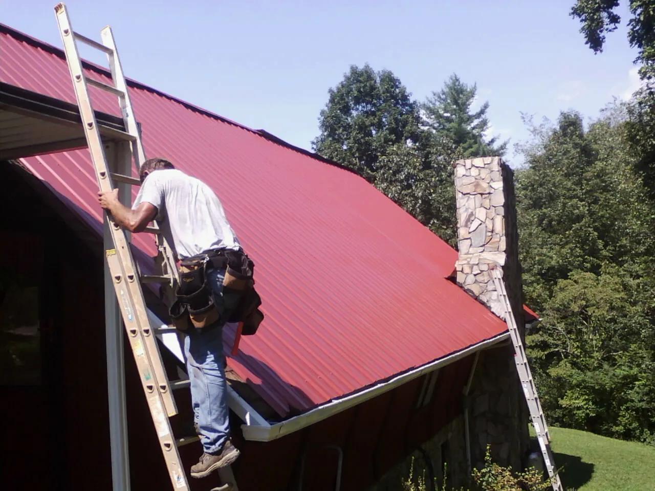 Roofer on a ladder beside a red metal roof and stone chimney — Soffit & Fascia Repair by Roofing Brothers in Astoria